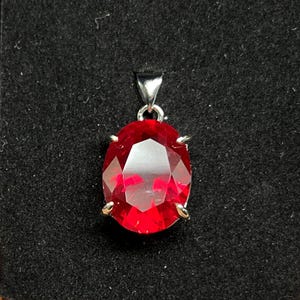 May include: A bright red oval gemstone pendant with a silver bail. The faceted stone is set in a silver metal setting. The pendant is displayed on a black surface.