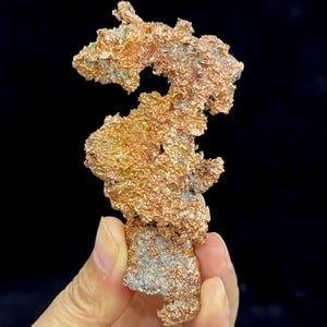 May include: A raw, metallic copper specimen featuring a jagged, irregular crystalline structure with a textured, reddish-brown surface. The mineral formation displays natural oxidation with small patches of green mineral deposits throughout the piece.