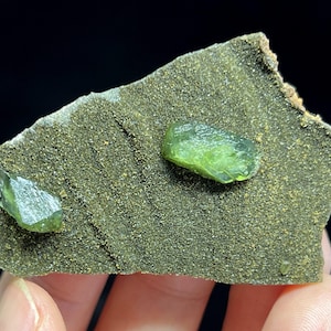 May include: A close-up of a mineral specimen featuring two vibrant, translucent green crystals embedded in a rough, dark green matrix. The crystals have a faceted appearance, and the matrix has a granular texture. The background is black.