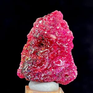 May include: A striking, deep red crystalline mineral specimen. The rough stone sits on a small, rounded white base, set against a dark backdrop. The mineral's surface reflects light, highlighting its natural texture and crystalline structure.