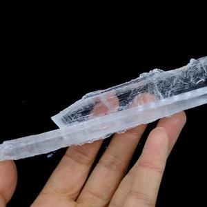 May include: A large, clear selenite crystal with a layered, striated texture. The crystal is held in a hand, showcasing its size and shape.
