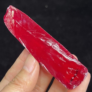 Può includere: Una gemma di rubino grezza, di un colore rosso intenso e profondo. La forma allungata presenta texture e imperfezioni naturali, che ne indicano lo stato non lucidato. La gemma è tenuta in mano su uno sfondo scuro.