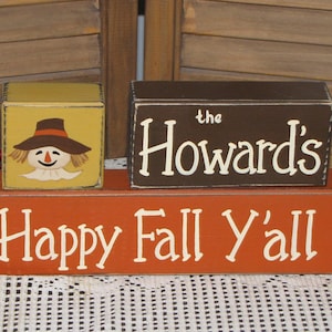 May include: Three wooden signs with a fall theme. The top sign is brown with white text that reads "the Howards". The middle sign is yellow with a painted scarecrow. The bottom sign is orange with white text that reads "Happy Fall Y'all".