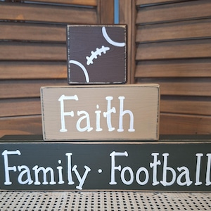 May include: Three wooden blocks stacked on top of each other. The top block is brown with a white football design. The middle block is tan with the word "Faith" in white. The bottom block is dark green with "Family Football" in white.