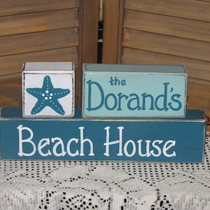 May include: A wooden sign with three blocks stacked on top of each other. The top block is light blue with white lettering that says "the Dorands". The middle block is white with a teal starfish. The bottom block is teal with white lettering that says "Beach House".