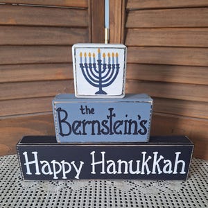 May include: Three wooden blocks with Hanukkah themes. The top block is white with a blue menorah design. The middle block is blue with "the Bernstein's" in black. The bottom block is navy blue with "Happy Hanukkah" in white.