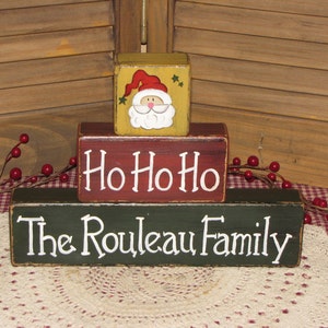 May include: A set of three wooden blocks stacked on top of each other. The top block is yellow with a painted image of Santa Claus. The middle block is red with the words "Ho Ho Ho" painted in white. The bottom block is green with the words "The Rouleau Family" painted in white.