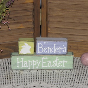 May include: Three wooden blocks stacked on top of each other. The top block is purple and says "The Bender's". The middle block is yellow and has a white bunny rabbit on it. The bottom block is green and says "Happy Easter".