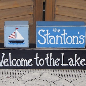 May include: A wooden sign with three blocks stacked on top of each other. The top block is light blue with white text that says "the Stantons". The middle block is light blue with a white sailboat with red and white stripes. The bottom block is black with white text that says "Welcome to the Lake".