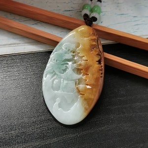 Könnte beinhalten: Eine Jade-Anhänger-Kette mit einem geschnitzten Design einer Figur in einer Landschaft. Der Anhänger ist oval geformt und hat ein braunes und grünes Farbschema. Die Kette besteht aus schwarzem Band und hat grüne Jadeperlen.