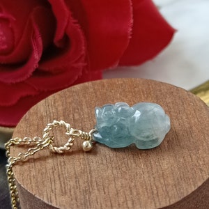 Könnte beinhalten: Ein Anhänger aus grünem Jade in Form eines hockenden Tieres, an einer goldenen Kette, auf einem Holzständer.