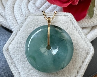 Puede incluir: Un collar con un colgante de jade redondo de color verde y una cadena de oro. El colgante está sobre un expositor hexagonal blanco.