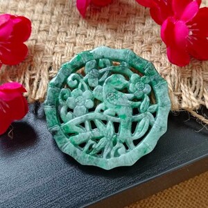 May include: A green jade pendant featuring a carved design of a bird and flowers. The pendant is round and has a delicate, intricate design.