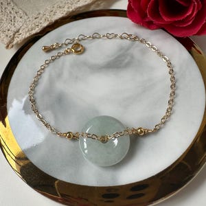 Könnte beinhalten: Ein zartes Armband mit goldfarbener Kette und einem hellgrünen Jade-Donut-Anhänger. Das Armband hat ein herzförmiges Kettendesign und wird auf einem weiß-goldenen Teller mit einer roten Rose im Hintergrund präsentiert.