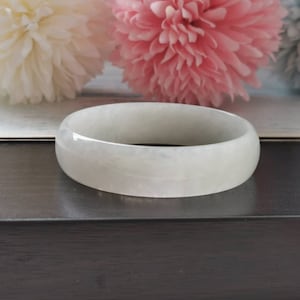Puede incluir: Un brazalete de jade blanco con un acabado liso y pulido. El brazalete se muestra sobre una superficie de madera oscura.