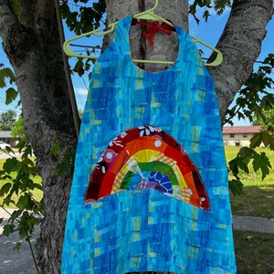 May include: A blue apron with a rainbow applique. The apron is made of blue fabric with a geometric pattern and a rainbow design in red, orange, yellow, green, and blue. It is hanging on a green hanger.