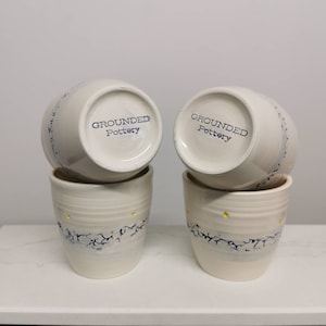 Può includere: Quattro tazze in ceramica bianca con un design blu e giallo. Le tazze sono impilate a coppie. Il fondo di ogni tazza ha le parole "Grounded Pottery" stampate.