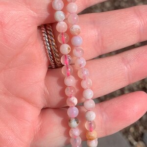 May include: Two delicate bracelets made of small, round, pink and white beads. The beads are strung together with a pink cord. The bracelets are held in a hand, with a silver ring visible on a finger. The beads have a slight variation in colour.