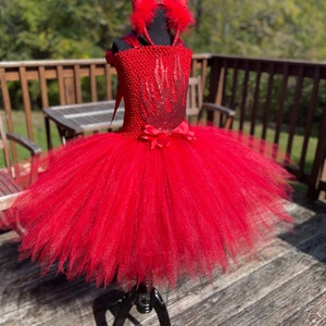 Könnte beinhalten: Roter Tüll-Tutu mit einem roten und schwarzen Flammenmuster auf dem Oberteil. Der Tutu steht auf einer Kleiderpuppe und hat eine rote Schleife um die Taille. Der Tutu ist mit roten Teufelshörnern mit schwarzen Federn verziert.