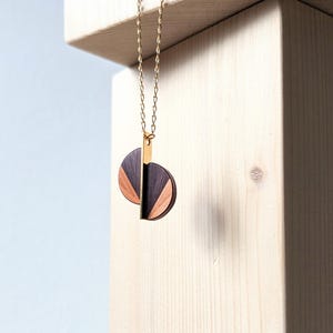 Könnte beinhalten: Eine goldene Kettenkette mit einem Anhänger aus zwei sich überlappenden Holzkreisen. Die Kreise sind aus dunklem Holz und hellem Holz gefertigt und bilden ein geometrisches Muster.
