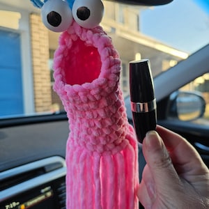 May include: A pink crocheted octopus car charm with googly eyes and blue pipe cleaner tentacles hanging from a rearview mirror. A hand is holding a black tube of lipstick.