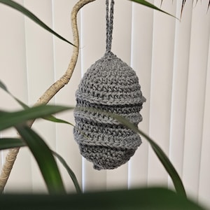 Pode incluir: Um vaso suspenso de crochê cinza, em forma de ovo, suspenso por um cordão cinza. A floreira tem um design em espiral ao redor do meio. O fundo inclui folhas verdes de plantas e uma persiana branca.