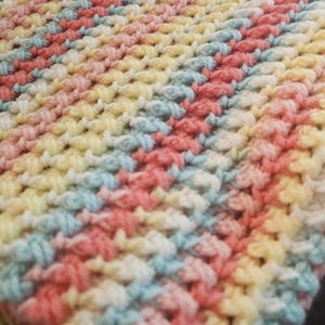 May include: A handmade crochet blanket in pastel colors. The blanket features rows of alternating colors including pink, yellow, blue, and white. The texture is dense and the stitches are clearly visible, creating a cozy and textured appearance.