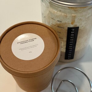 May include: A close-up of sourdough baking supplies. A brown paper container with a label that reads "SOURDOUGH STARTER FEEDING FLOUR" sits next to a glass jar with flour and a metal whisk. The jar has temperature markings.