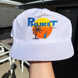 May include: A white baseball cap featuring a graphic print of a yellow and orange sun, a black palm tree silhouette, and a small blue sailboat. The text PHUKET is printed in large blue letters above the sun, with the word Thailand in smaller black text.