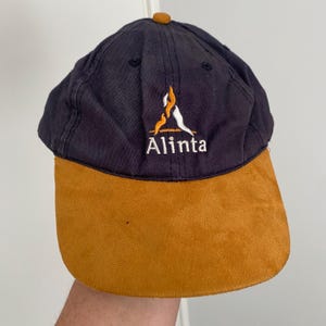May include: A navy blue baseball cap with a tan suede-like brim and a small tan button on top. The front features an embroidered logo with an orange and white flame-like graphic above the white text Alinta, centered on the crown.