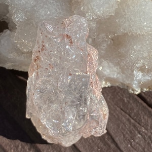 May include: A clear, translucent crystal with a pink hue and a rough, textured surface. The crystal has a natural, organic shape and is likely a piece of rose quartz.
