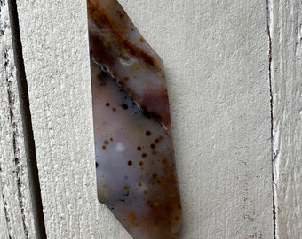 May include: A polished, elongated gemstone with a unique pattern of brown, white, and reddish-purple hues. The stone has a smooth, glossy surface and a slightly irregular shape. Small, dark brown spots are scattered across the surface. The gemstone is set against a white wooden background.