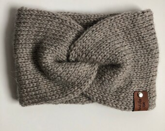 May include: A gray knitted headband with a twisted knot design. The headband is made of a thick, textured yarn and features a small brown leather tag with text. The accessory is designed to keep the ears warm.