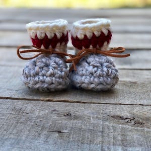 May include: A pair of handmade baby booties. The booties are crocheted with gray yarn for the foot and white and red yarn for the cuff. Brown leather ties secure the booties. The background is a weathered wooden surface.