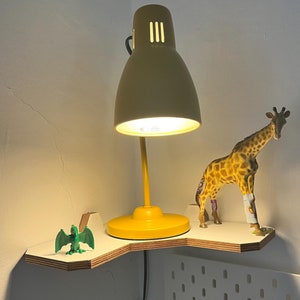 May include: A yellow desk lamp with a white shade sits on a white corner shelf. A green dinosaur toy and a brown giraffe toy are also on the shelf.