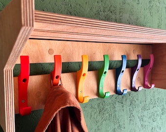 May include: A wooden coat rack for the wall, featuring seven brightly coloured hooks. The hooks are red, orange, yellow, green, blue, purple, and pink. A brown garment hangs from one of the hooks. The rack is made of light-coloured wood.