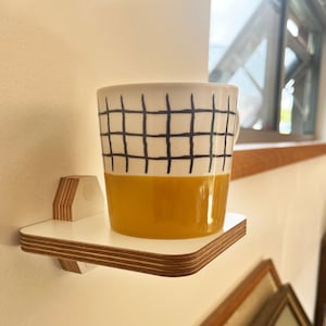 May include: A small, white ceramic mug with a black grid pattern and a yellow glaze on the bottom half. The mug is sitting on a small, white shelf attached to a wall.