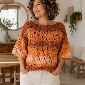 Puede incluir: Top de ganchillo en tonos naranja, melocotón y marrón. La parte superior de manga corta tiene un cuello barco y un tejido abierto. La modelo lleva la parte superior con pantalones claros.