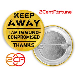May include: Yellow button with black text that reads "Keep Away I Am Immuno-Compromised Thanks". The button has a silver back with a pin.