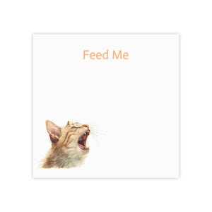 以下が含まれることがあります： オレンジ色の「Feed Me」というテキストが入った正方形の白いメモ帳。あくびをしているトラ猫の水彩イラストが左下にあります。猫は茶色とタン色の毛皮をしています。