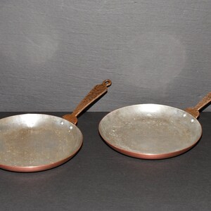 May include: Two copper-bottomed pans with hammered bronze handles. The pans have a shallow, round cooking surface and a long handle with a loop for hanging. The metal has a rustic, aged appearance, suggesting they are vintage or handcrafted.