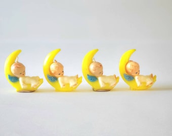 May include: Four vintage baby-themed candles. Each candle features a sleeping baby figure in a cream color, resting on a yellow crescent moon. The baby has small blue wings. The candles are arranged in a row against a white background.