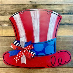 May include: A wooden Uncle Sam hat decoration in red, white, and blue. The hat has red and white stripes, a blue band with white polka dots, and a red brim. A decorative bow with red, white, and blue ribbons adds detail. A wire loop is attached.