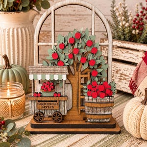 May include: A layered wooden decorative sign featuring an arched window frame, an apple tree with red fruit, a small cart labeled FARM FRESH APPLES, and a wooden barrel filled with red apples, all mounted on a flat rectangular base.