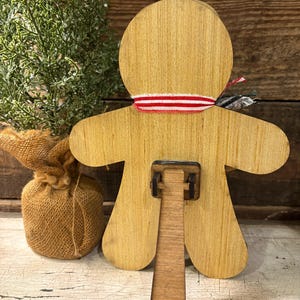 May include: Wooden gingerbread man-shaped decoration with a red and white striped ribbon around the neck. The back features a stand for display. A small burlap sack and a sprig of greenery are in the background.