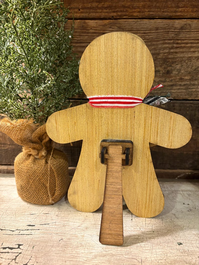 May include: Wooden gingerbread man-shaped decoration with a red and white striped ribbon around the neck. The back features a stand for display. A small burlap sack and a sprig of greenery are in the background.