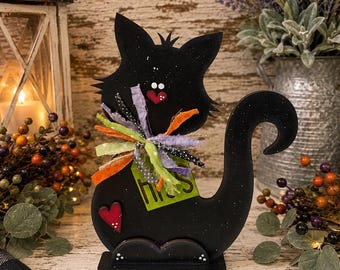 May include: A black wooden cat cutout featuring a speckled finish, a red heart-shaped nose, and a small red heart on its body. It wears a multi-colored fabric ribbon collar and holds a lime green tag with the text "his" in dark brown letters.