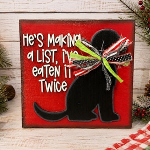 May include: A square red wooden sign featuring a black silhouette of a dog wearing a multi-colored fabric bow. The sign displays white text that reads, "He's Making a LIST, i've eaten it Twice," with a distressed finish on the edges of the board.