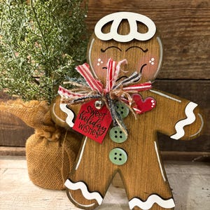 May include: Wooden gingerbread man Christmas decoration with white icing details, green button accents, and a red tag that says "Sweet holiday wishes." The gingerbread man has a red heart and is decorated with a red and white striped bow.