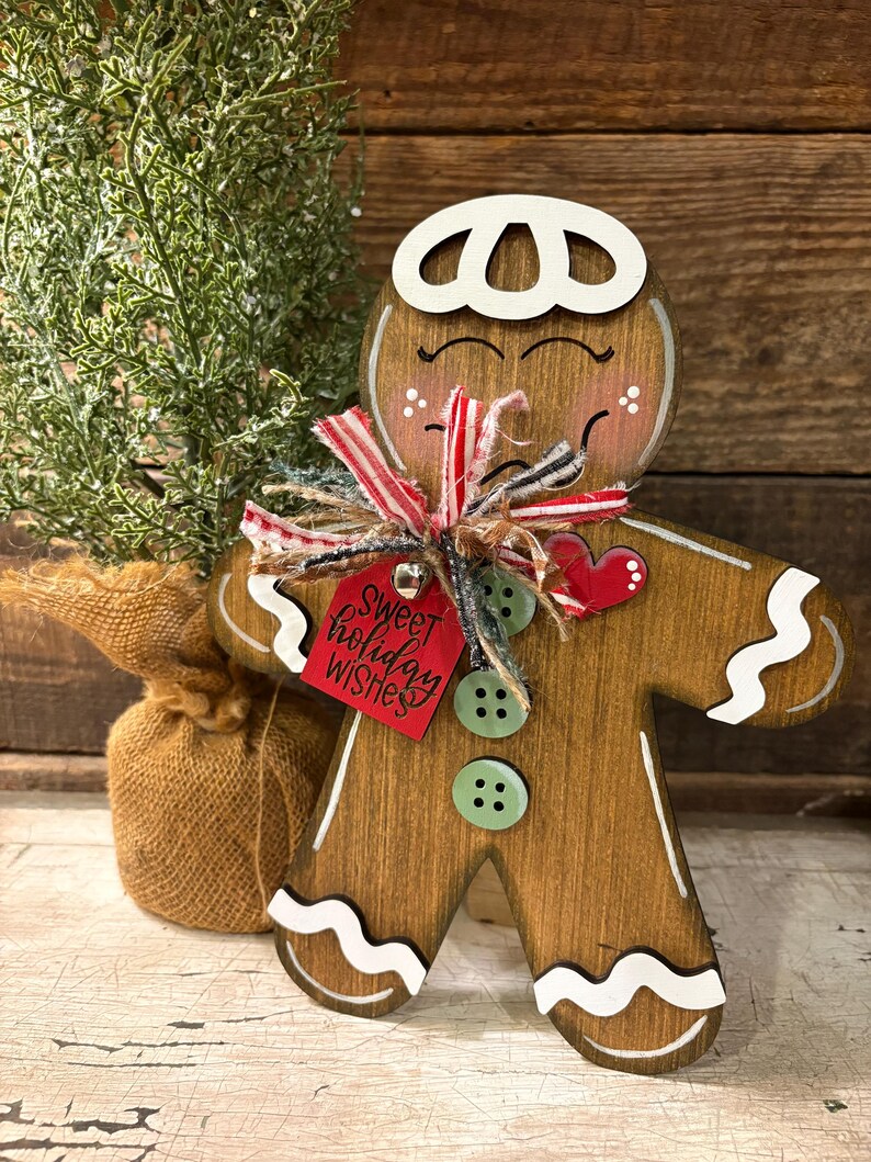 May include: Wooden gingerbread man Christmas decoration with white icing details, green button accents, and a red tag that says "Sweet holiday wishes." The gingerbread man has a red heart and is decorated with a red and white striped bow.